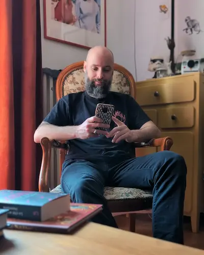 Homme barbu portant un t-shirt noir et un jean assis sur un fauteuil et regardant son téléphone. 