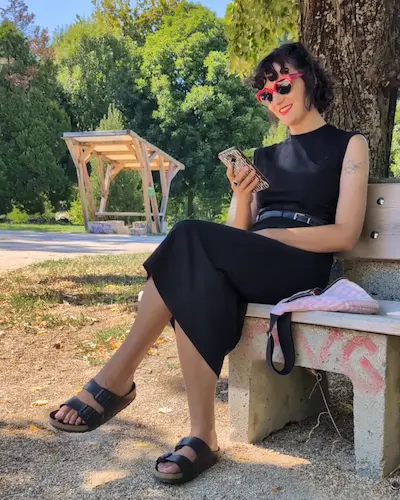 Une femme portant une robe noire assise sur un banc dans un parc, regardant en souriant son smartphone.