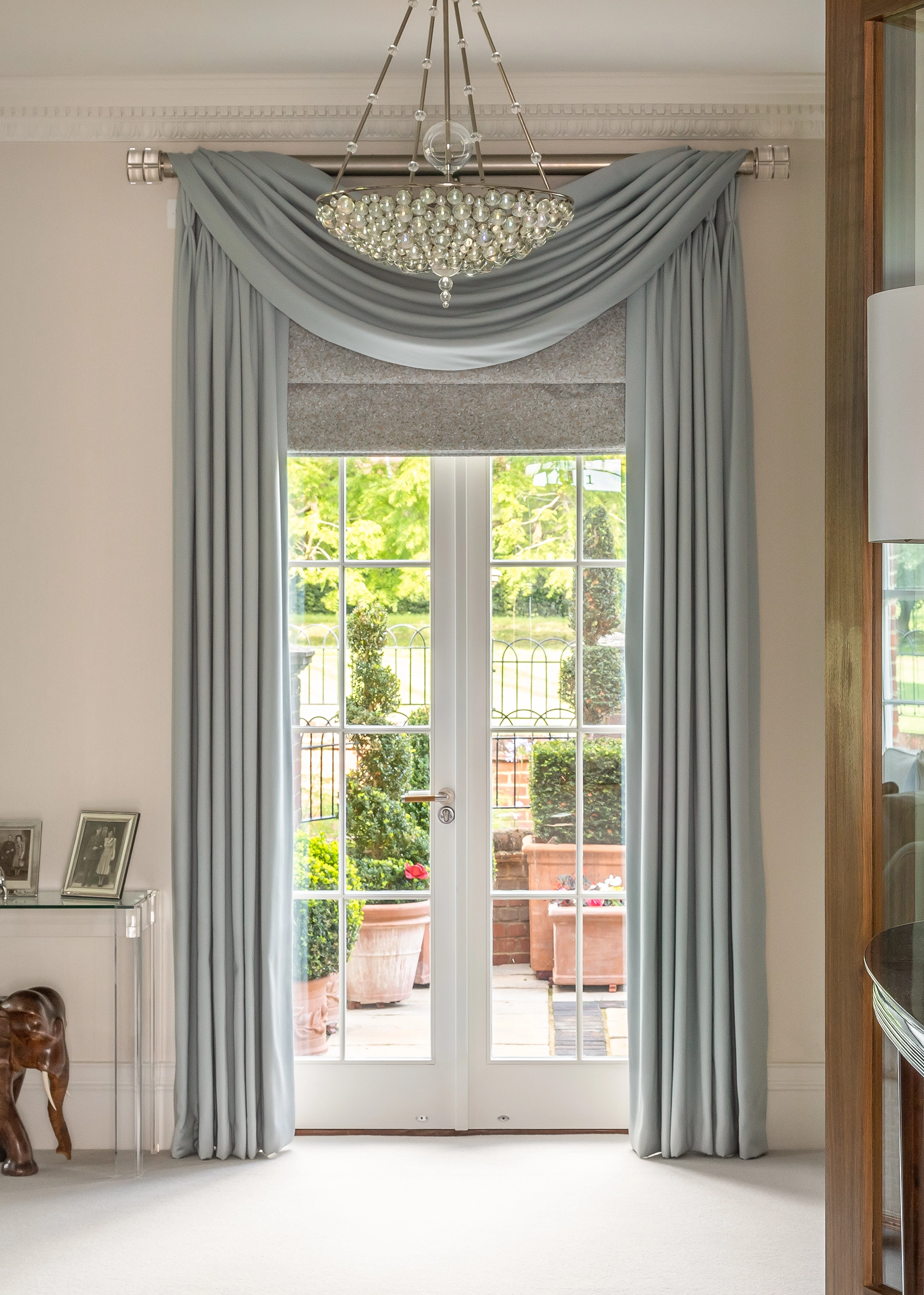 Pale blue silk curtains with a swagged pelmet framing French doors onto a garden terrace, beneath a crystal chandelier at Mia, London