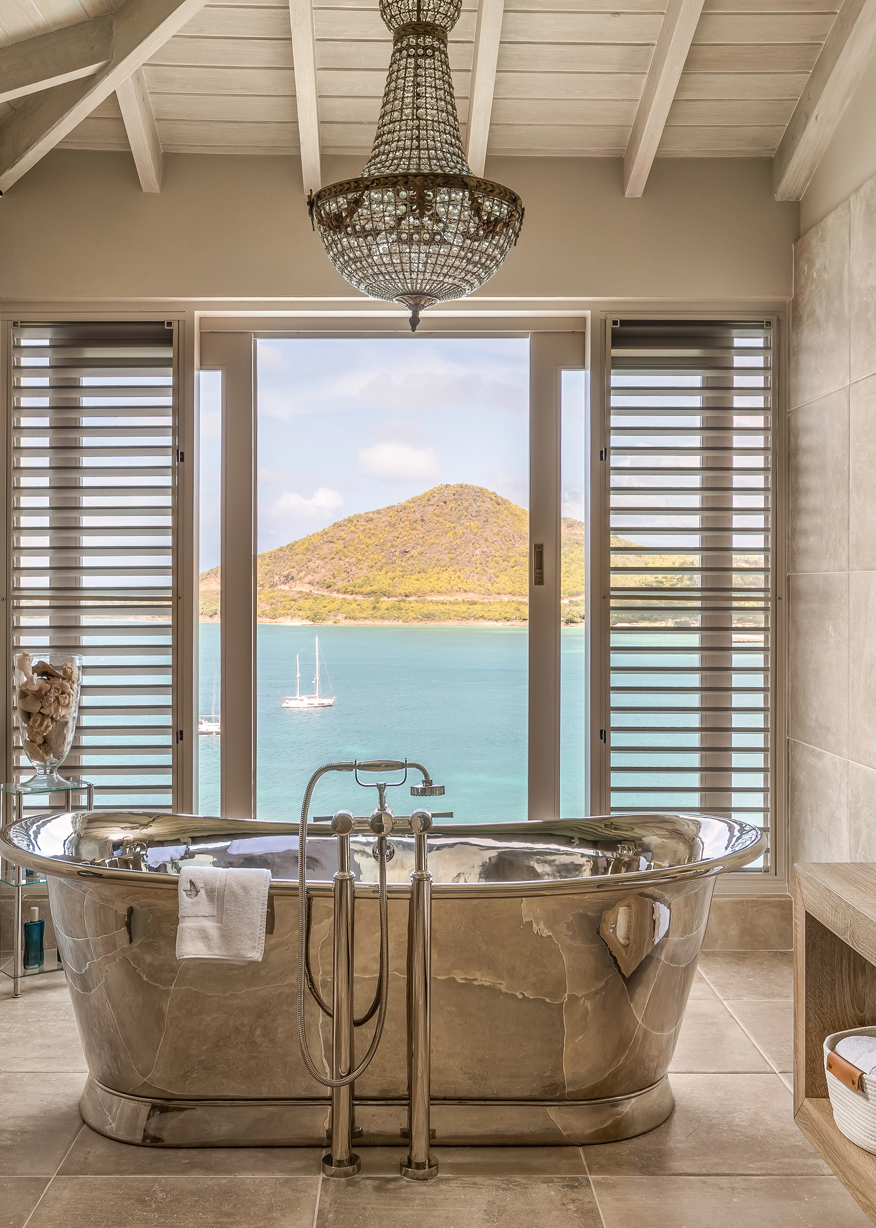 Freestanding polished nickel bathtub beneath a crystal chandelier, with shuttered windows framing a sea and hillside view at Villa Papillon, Antigua