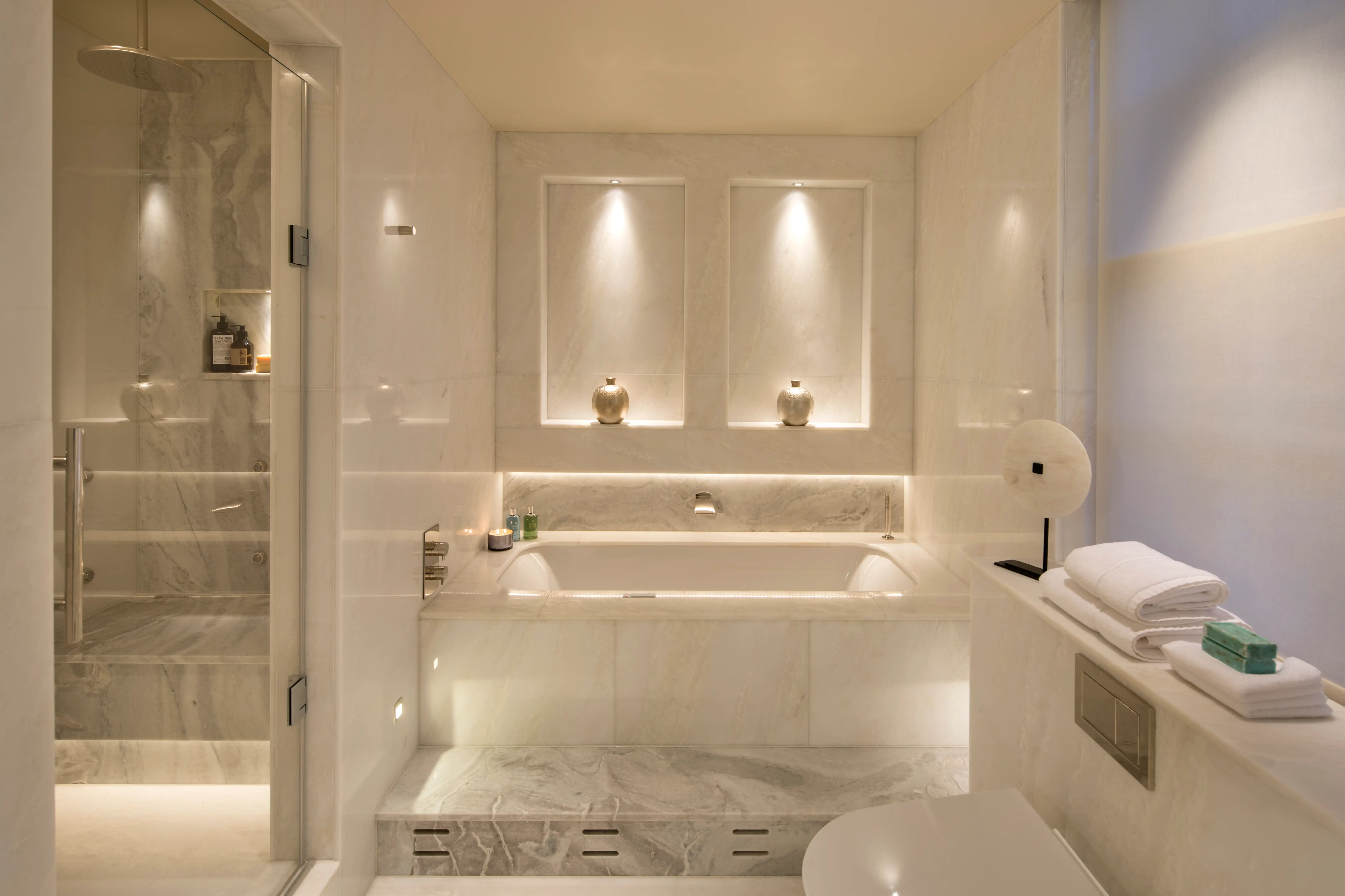 White marble bathroom with a built-in bath, softly lit wall niches and a walk-in glass shower in a Mayfair home by Ward & Co