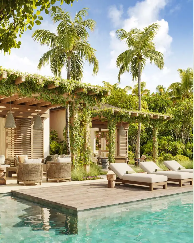 Sun loungers on a timber pool deck beside a vine-covered pergola with rattan seating and palm trees at a Caribbean resort
