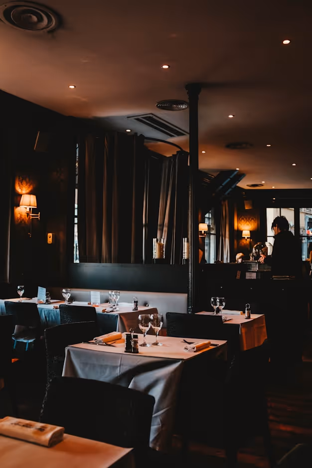Dimly lit restaurant interior with tables set with wine glasses, napkins, and condiments, and a person standing near the counter.