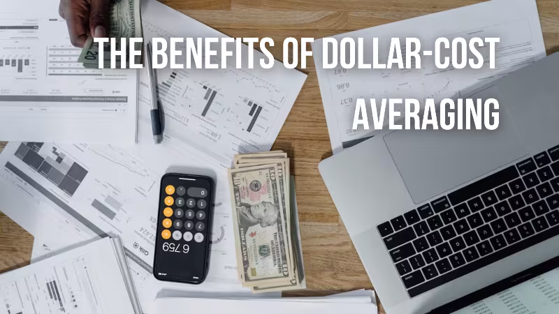 A top-down view of a desk with financial documents, a calculator, a stack of dollar bills, and a laptop. A hand is holding some money in the upper left corner. The text overlay reads: "THE BENEFITS OF DOLLAR-COST AVERAGING
