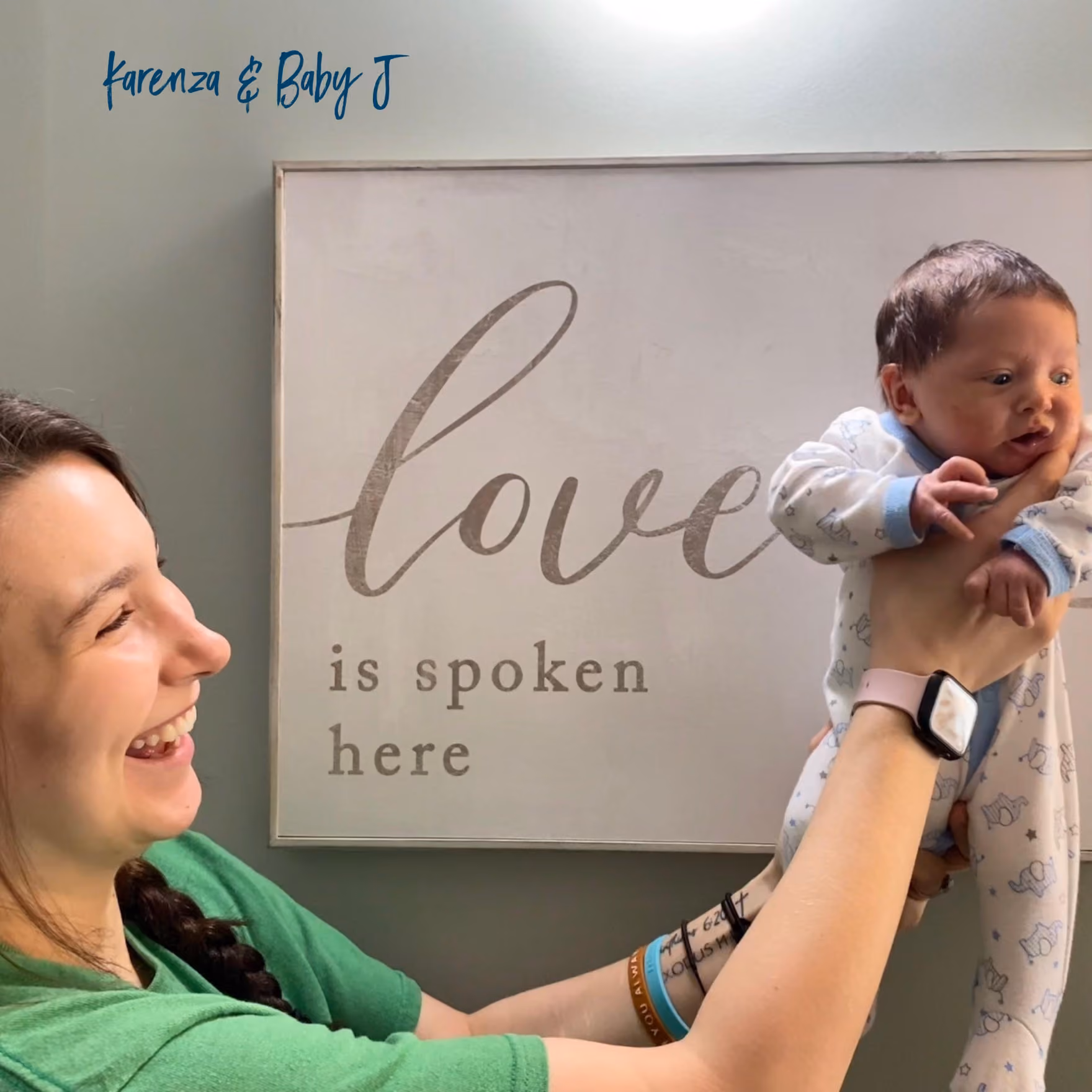 Woman in green shirt laughing and holding up a baby in white pajamas with blue trim, standing in front of a wall sign that says 'love is spoken here.'