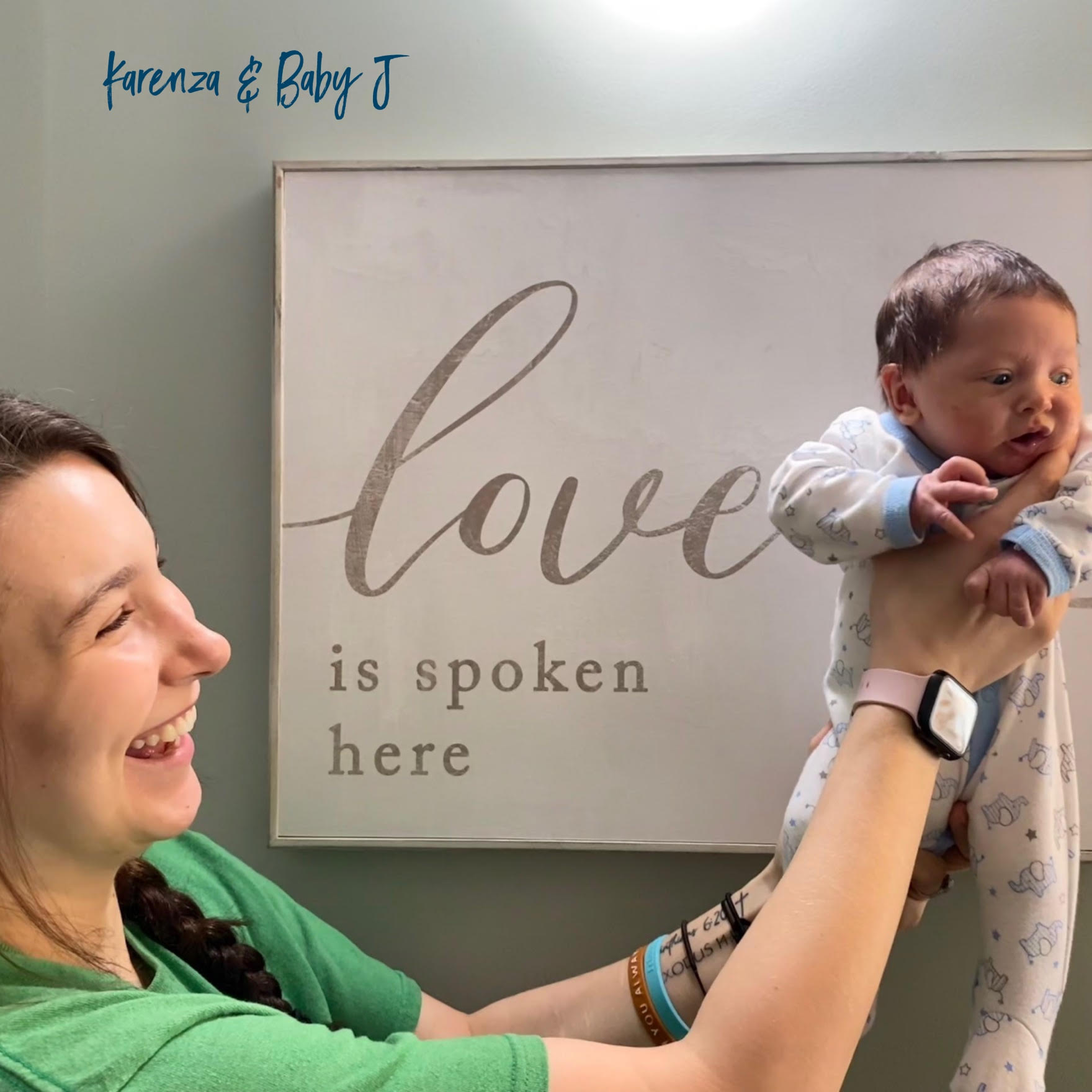 Woman in green shirt laughing and holding up a baby in white pajamas with blue trim, standing in front of a wall sign that says 'love is spoken here.'