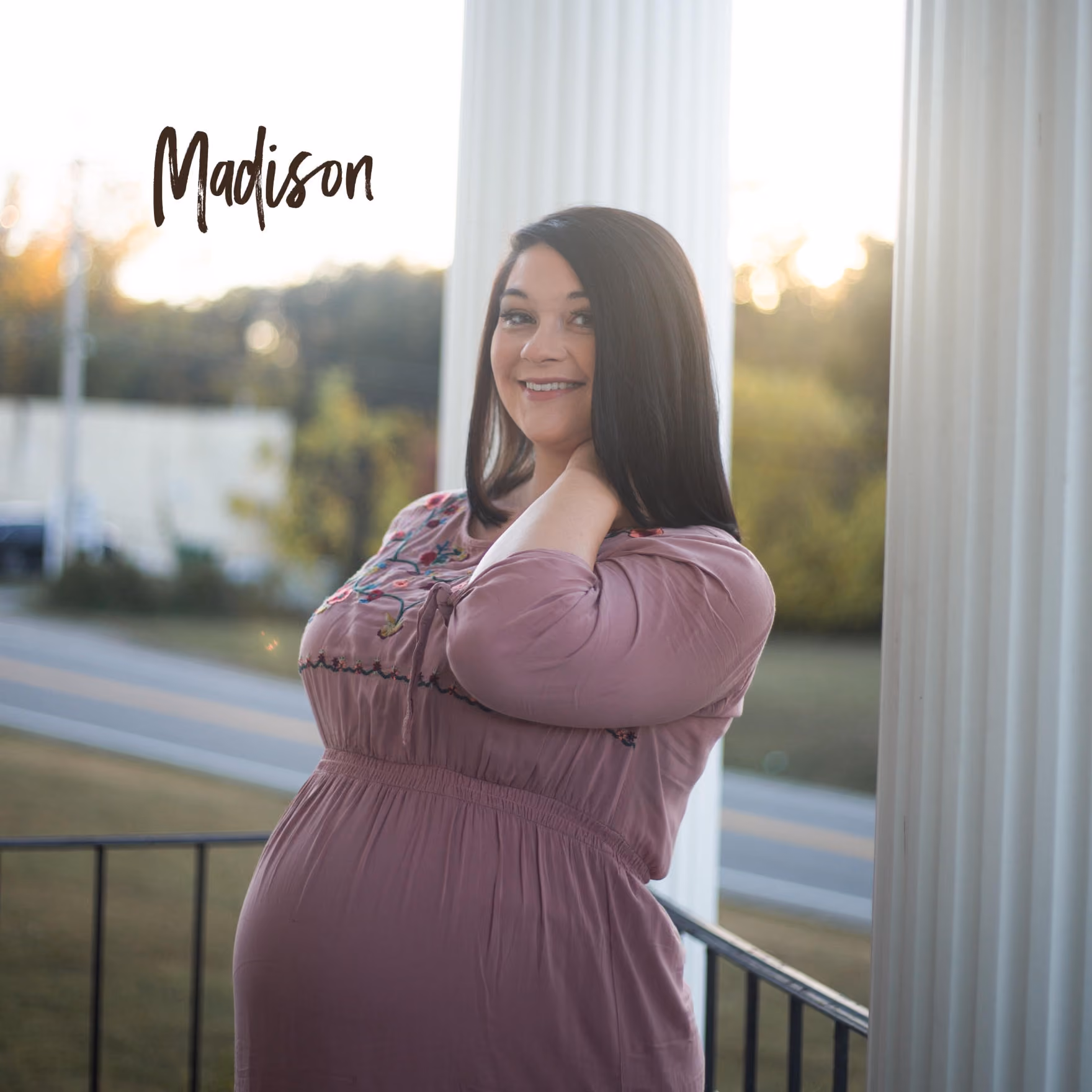 Smiling pregnant woman with long dark hair in a mauve dress standing outdoors near white columns at sunset.
