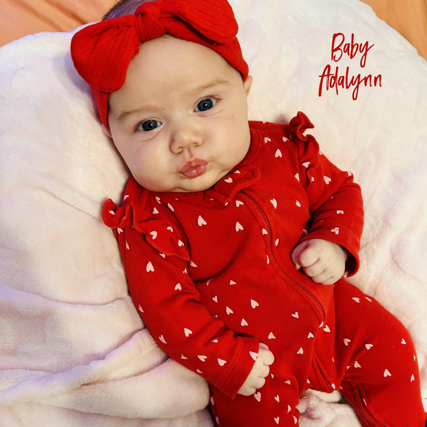 Baby wearing a red outfit with white hearts and a red headband lying on a soft white blanket.