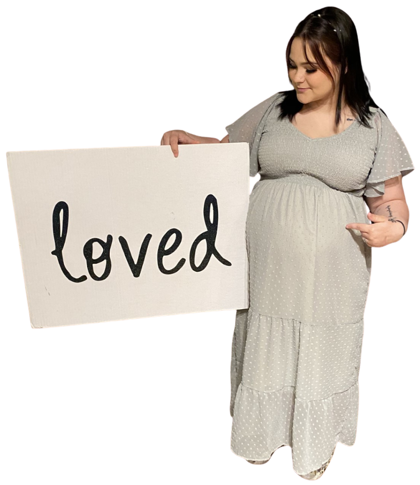 Pregnant woman in a light-colored dress holding a sign that says 'loved' while pointing at her belly.