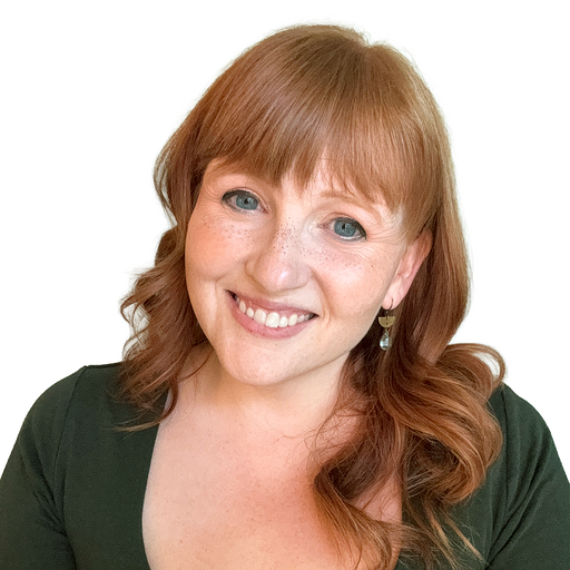 Smiling red-haired woman with freckles wearing a dark green top and dangling earrings on a white background.