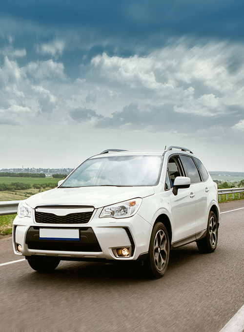 White SUV driving on a highway with green fields and cloudy sky in the background.