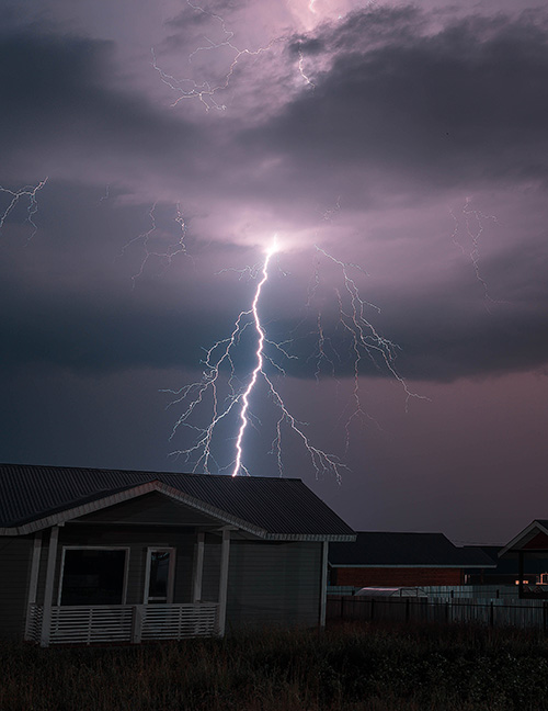 A house sits in a rural area with a stormy backdrop and a large lightning bolt shooting through the sky.