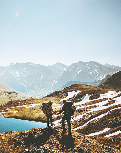 Two hikers holding hands on a mountain ridge with patches of snow and a blue lake below, surrounded by distant peaks under a clear sky.