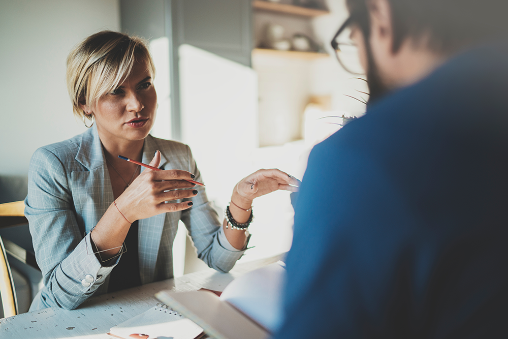 Two people having a focused discussion, one woman holding a pencil and gesturing with her hand across the table.