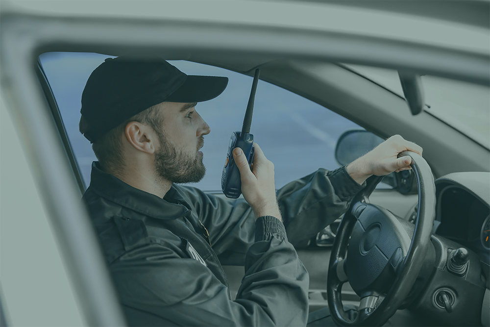 Security guard in uniform speaking into a walkie-talkie while driving a car.