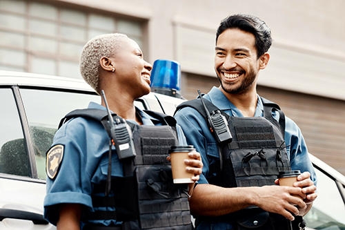 Two police officers smiling and holding coffee cups while standing in front of a police car.