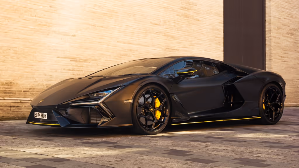 A matte black Lamborghini Revuelto hybrid supercar, featuring yellow accents on the wheels and body, parked against a light-coloured brick wall.