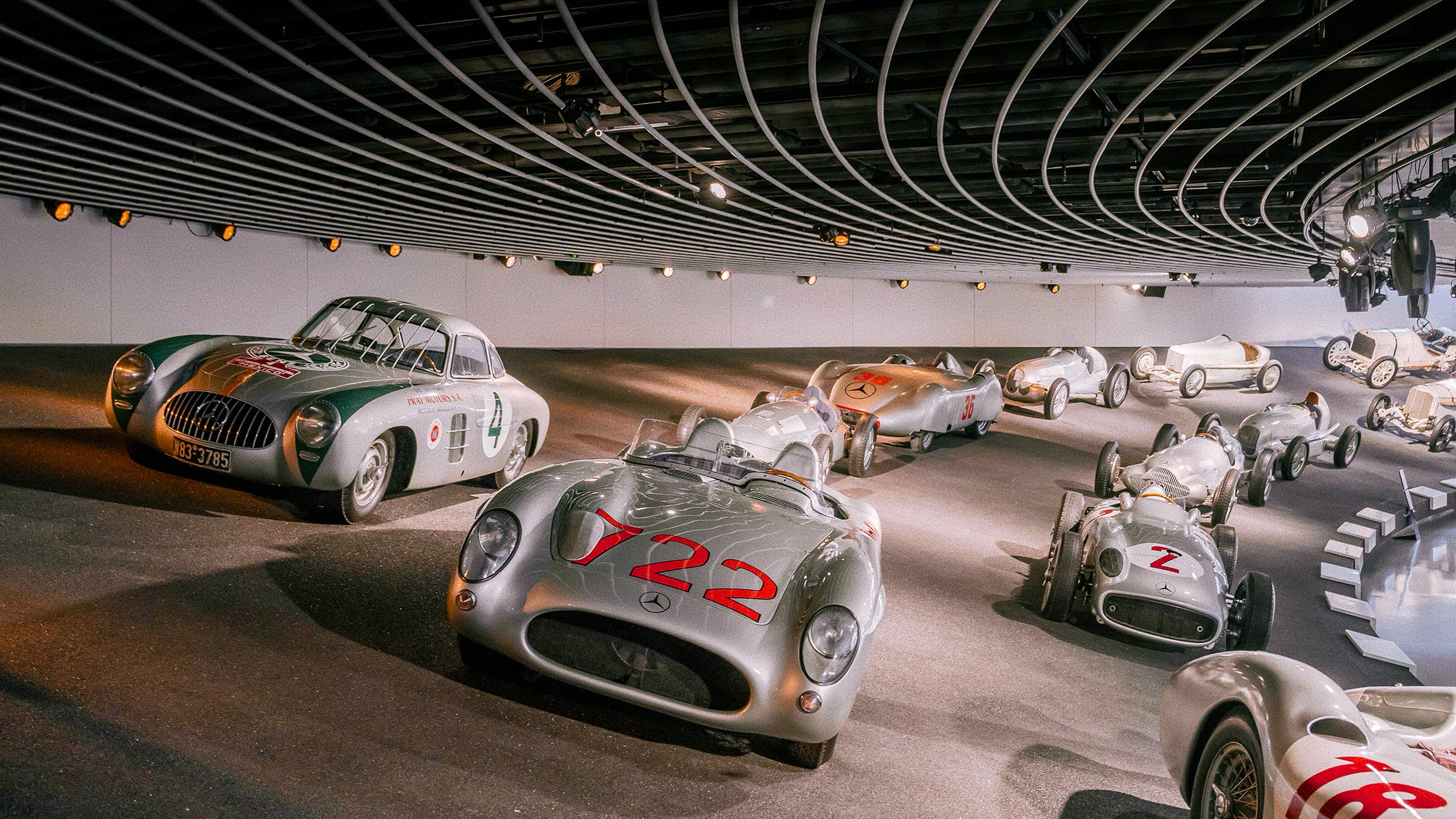 A display of classic silver Mercedes-Benz race cars and sports cars, including the 300 SL Gullwing and open-top racers, inside a modern, curved museum hall.