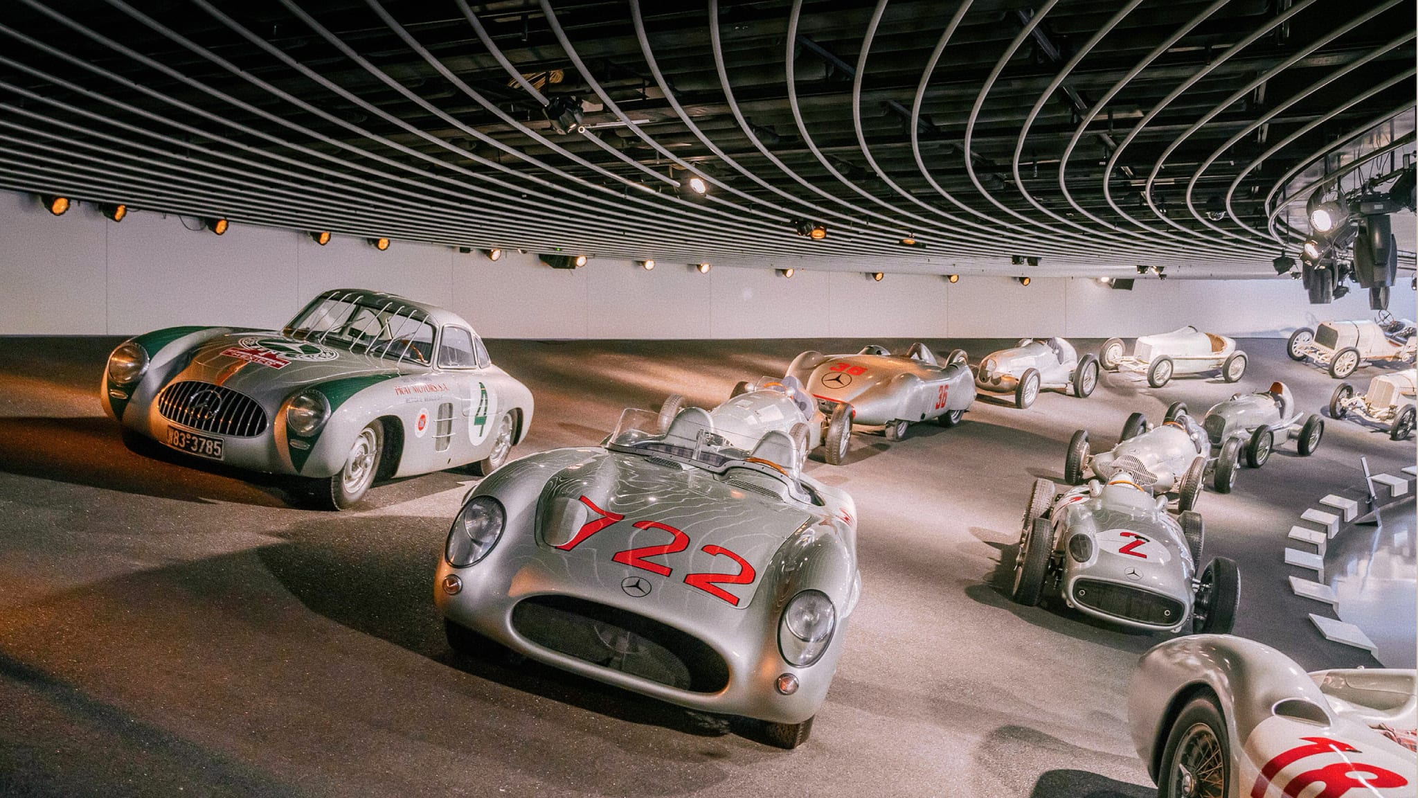 A display of classic silver Mercedes-Benz race cars and sports cars, including the 300 SL Gullwing and open-top racers, inside a modern, curved museum hall.