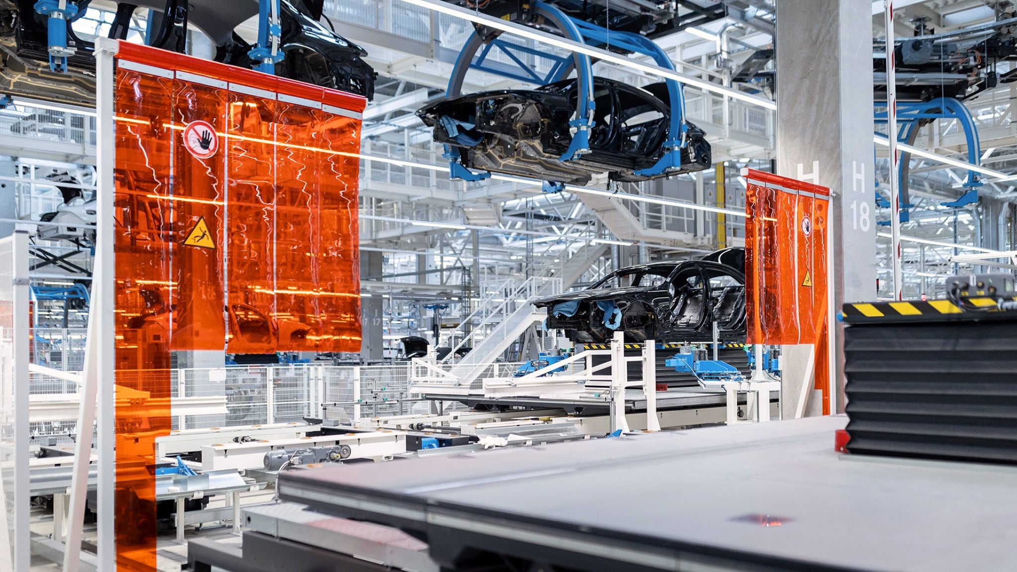 An automated automotive production line in the Mercedes-Benz Factory 56, showing car bodies being moved by overhead conveyors and precision machinery, with orange safety screens.