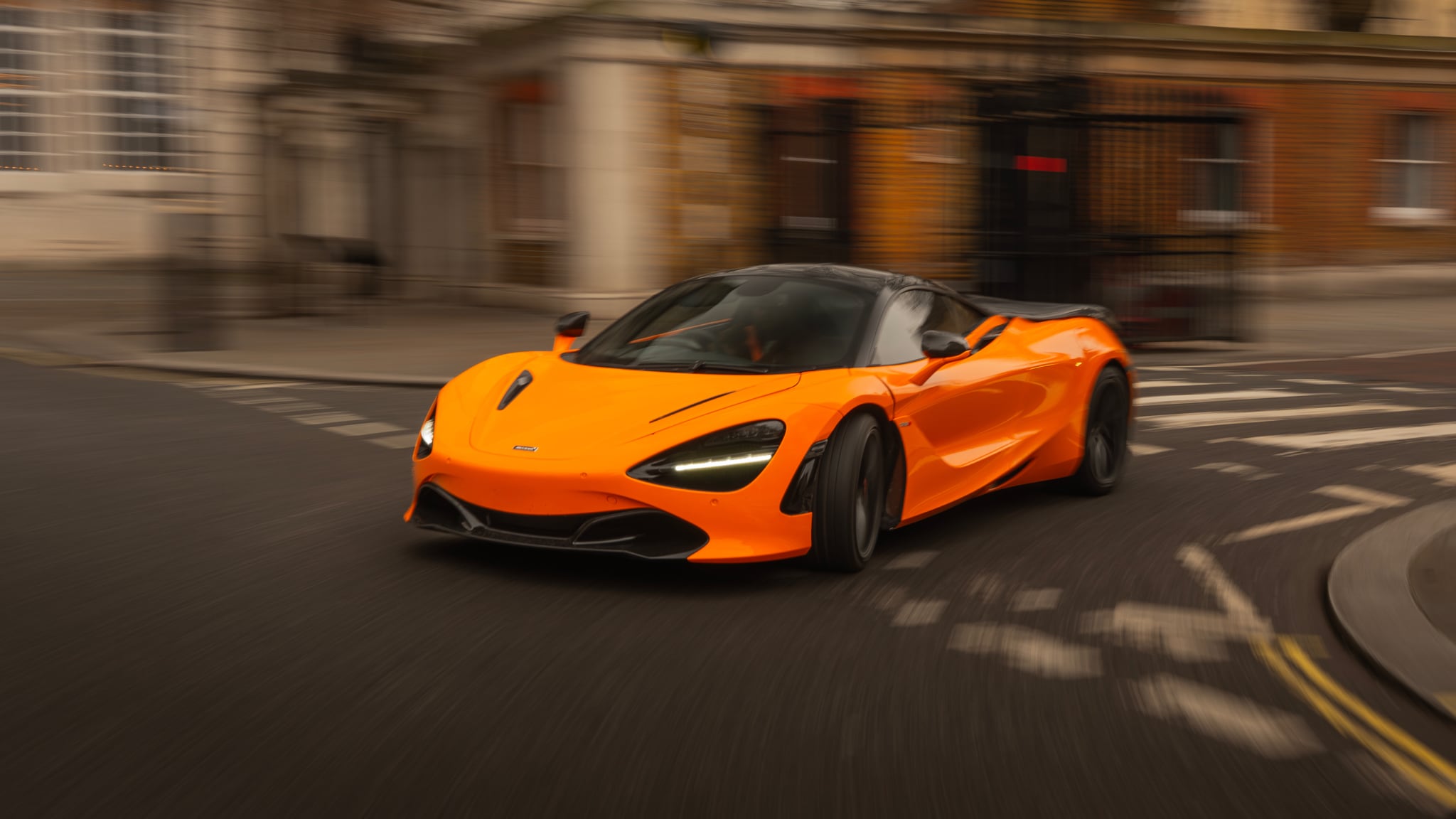An orange McLaren 720S supercar moving quickly on a city street, with the background blurred to emphasise speed.