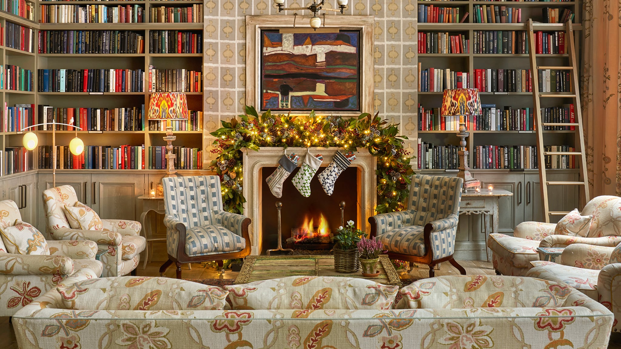 A cozy library/lounge area with floor-to-ceiling bookshelves, comfortable patterned seating, a fireplace with Christmas stockings, and a lit garland.