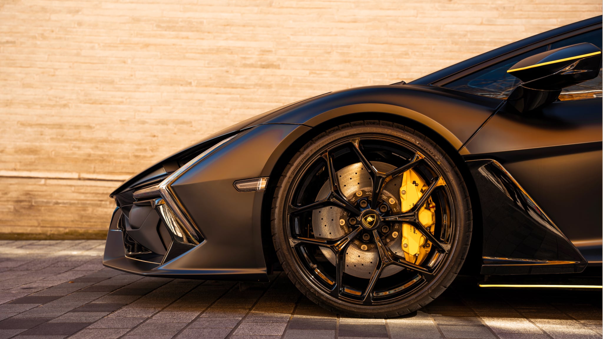A low-angle close-up of a black Lamborghini Revuelto's front quarter panel and wheel, featuring a multi-spoke black rim, a large drilled brake rotor, and a bright yellow brake caliper, set against a pale stone wall.