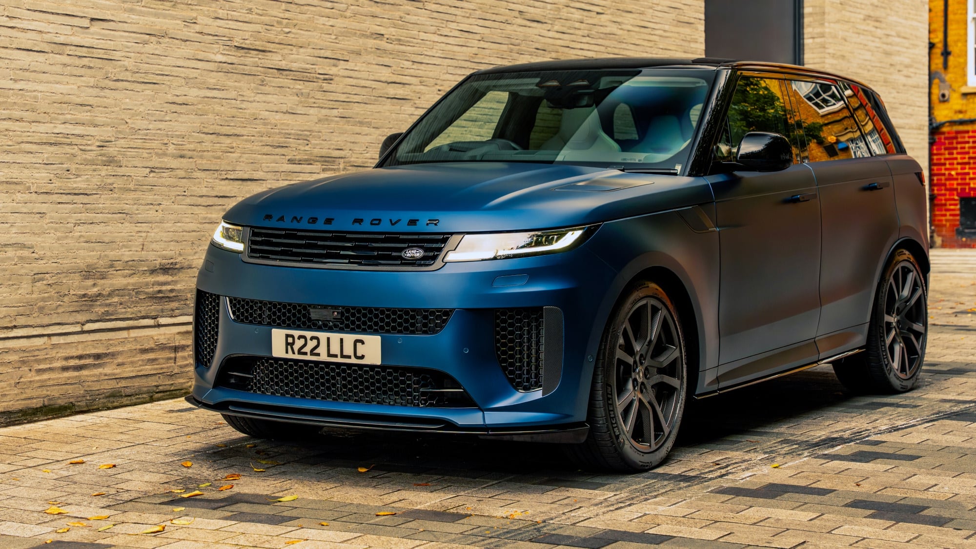 A low-angle front three-quarter view showcasing the matte blue Range Rover Sport SV's aggressive styling and black alloy wheels, parked against a modern brick facade.