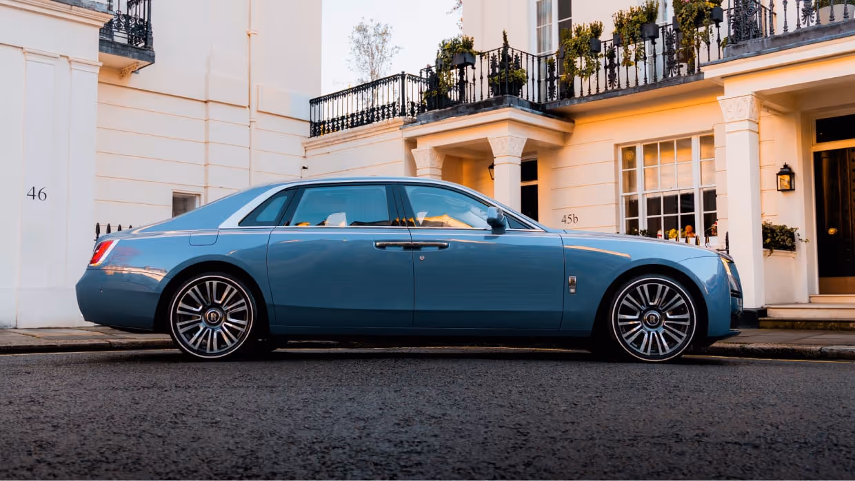 The Rolls-Royce Ghost in bespoke light blue/grey, parked elegantly outside a classic white stucco London residence, emphasizing LLC's discreet luxury chauffeur service.