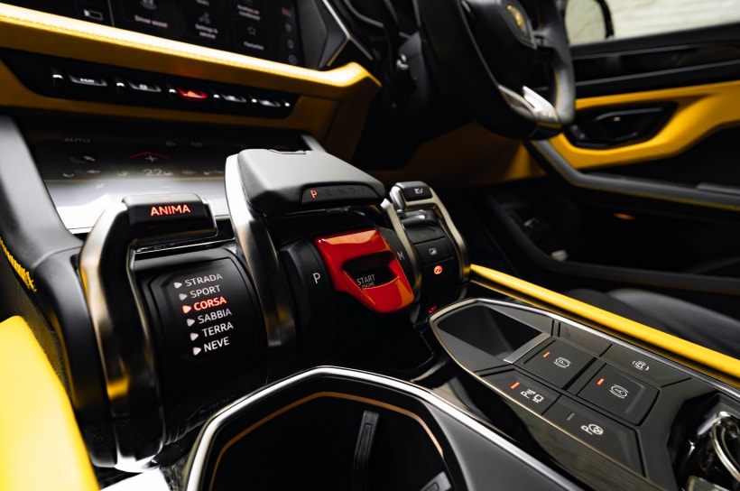 A close-up of the Lamborghini Urus SE center console showing the "Tamburo" selector set to Corsa mode, with a red flip-up start button and yellow leather trim.