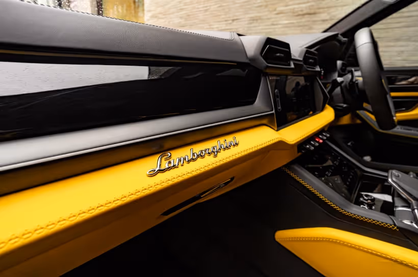 A close-up of the passenger side dashboard in a Lamborghini Urus, featuring yellow leather with contrast stitching, a chrome Lamborghini script logo, and carbon fiber accents.