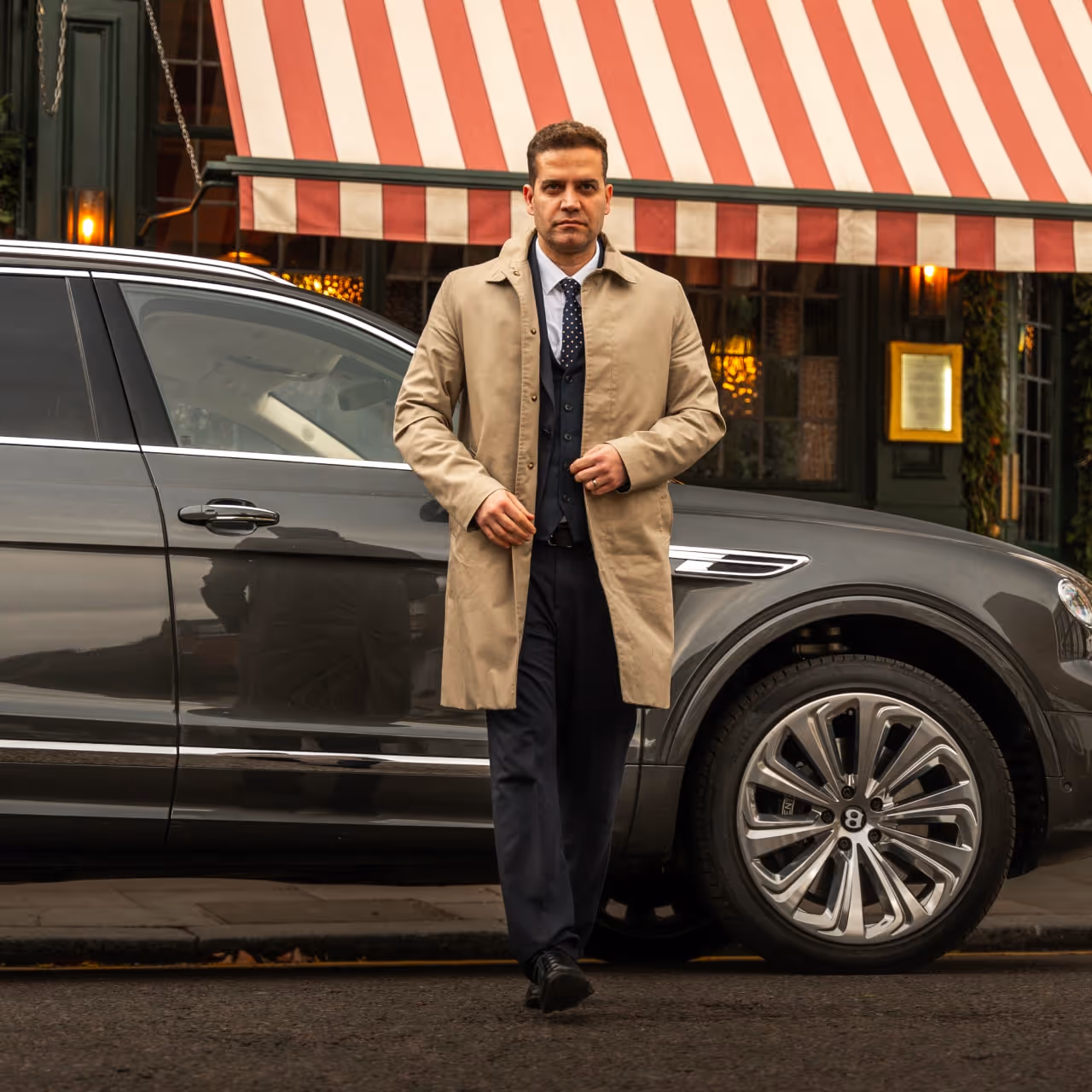 A professional chauffeur in a trench coat standing beside a luxury executive vehicle, representing bespoke long-term chauffeur solutions for private households and VIPs.