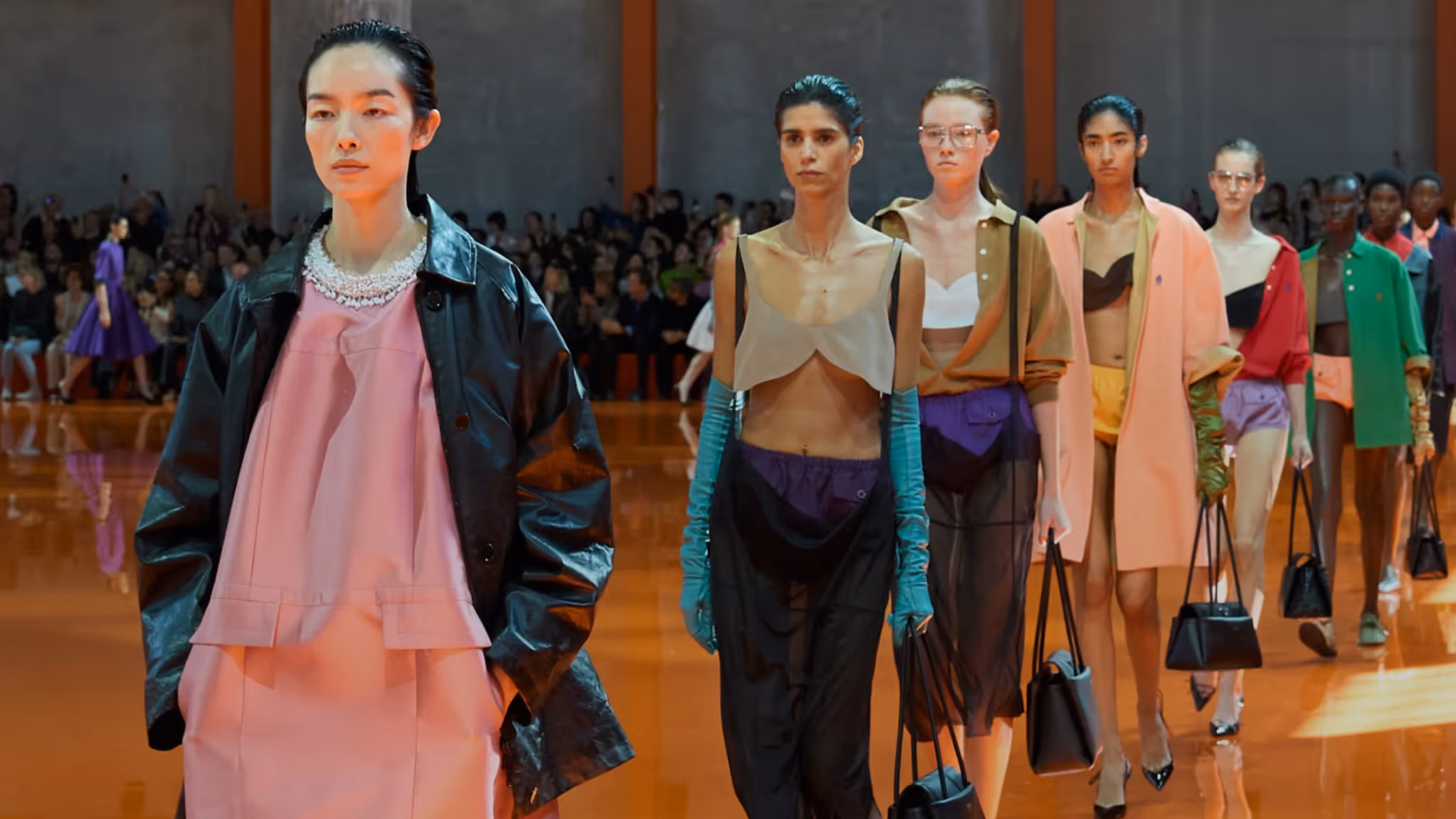 Models walking a runway during a high-fashion show featuring a layered and structured designer outfits in bold colors, set against a glossy orange floor and industrial backdrop, with an audience watching in the background.