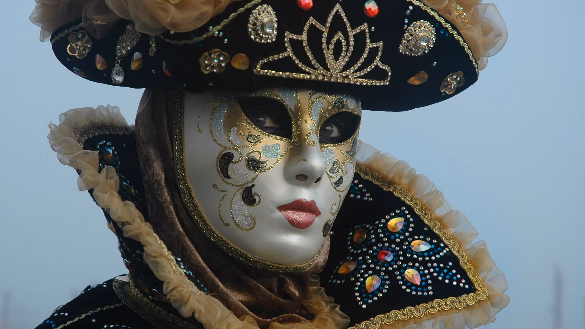 Close-up of a person wearing an ornate Venetian Carnival mask with gold detailing, a feathered hat and historical costume elements.
