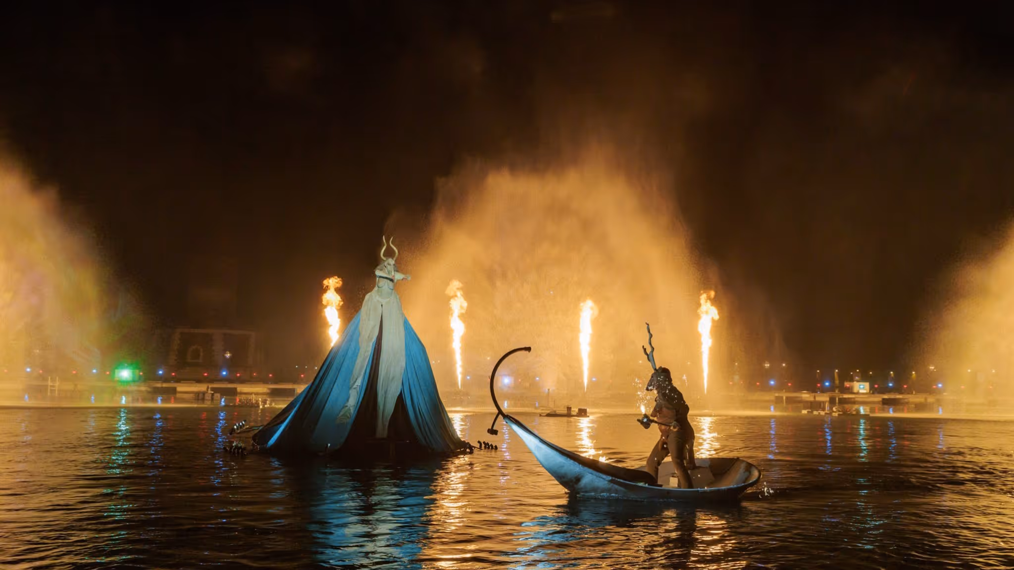 Night-time Carnival performance on the water featuring a costumed figure in a boat and a large illuminated installation inspired by mythological themes.