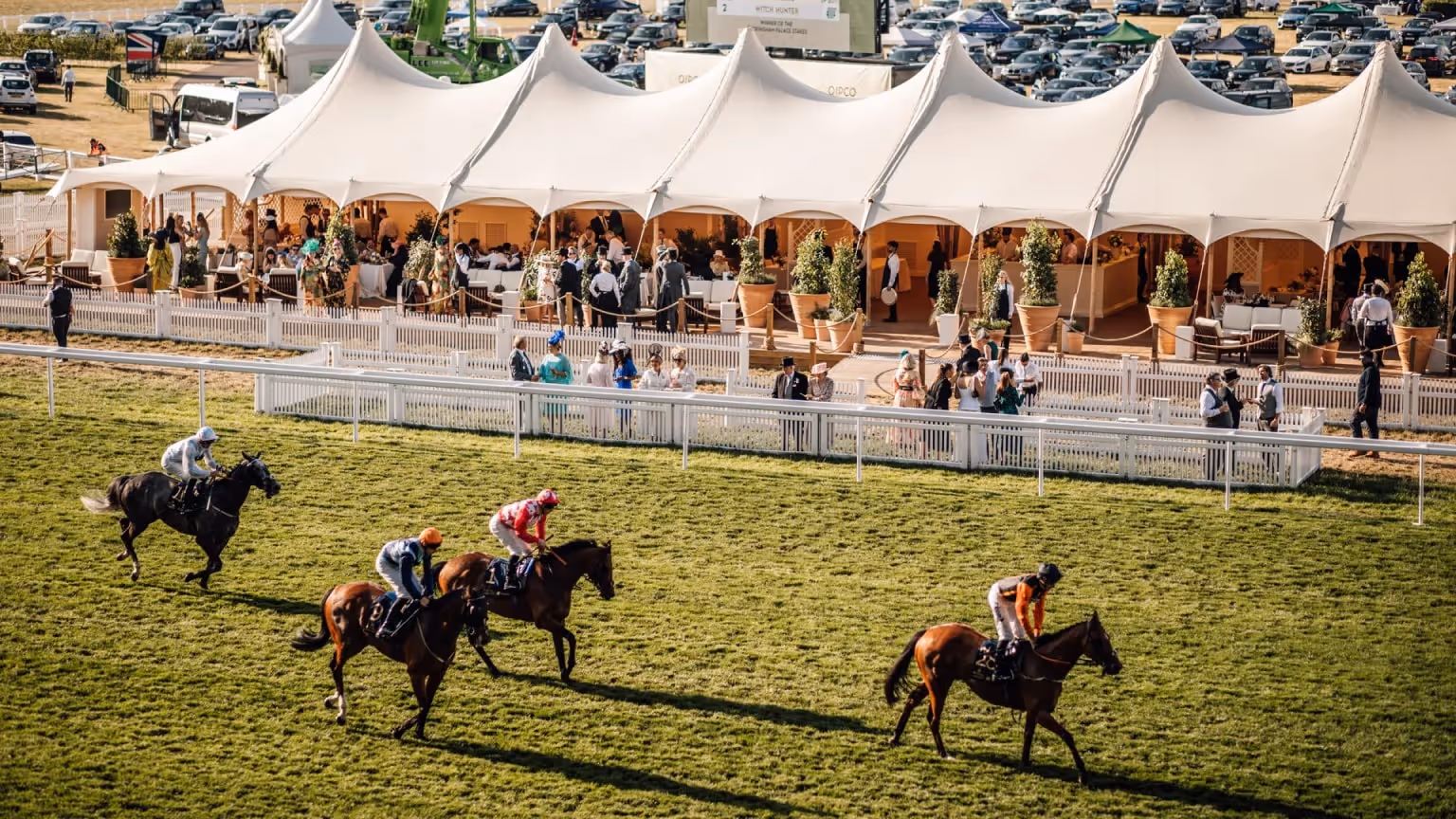 Two racehorses with jockeys in coloured silks gallop along a grass track in front of a white hospitality marquee with seated spectators behind a railing.
