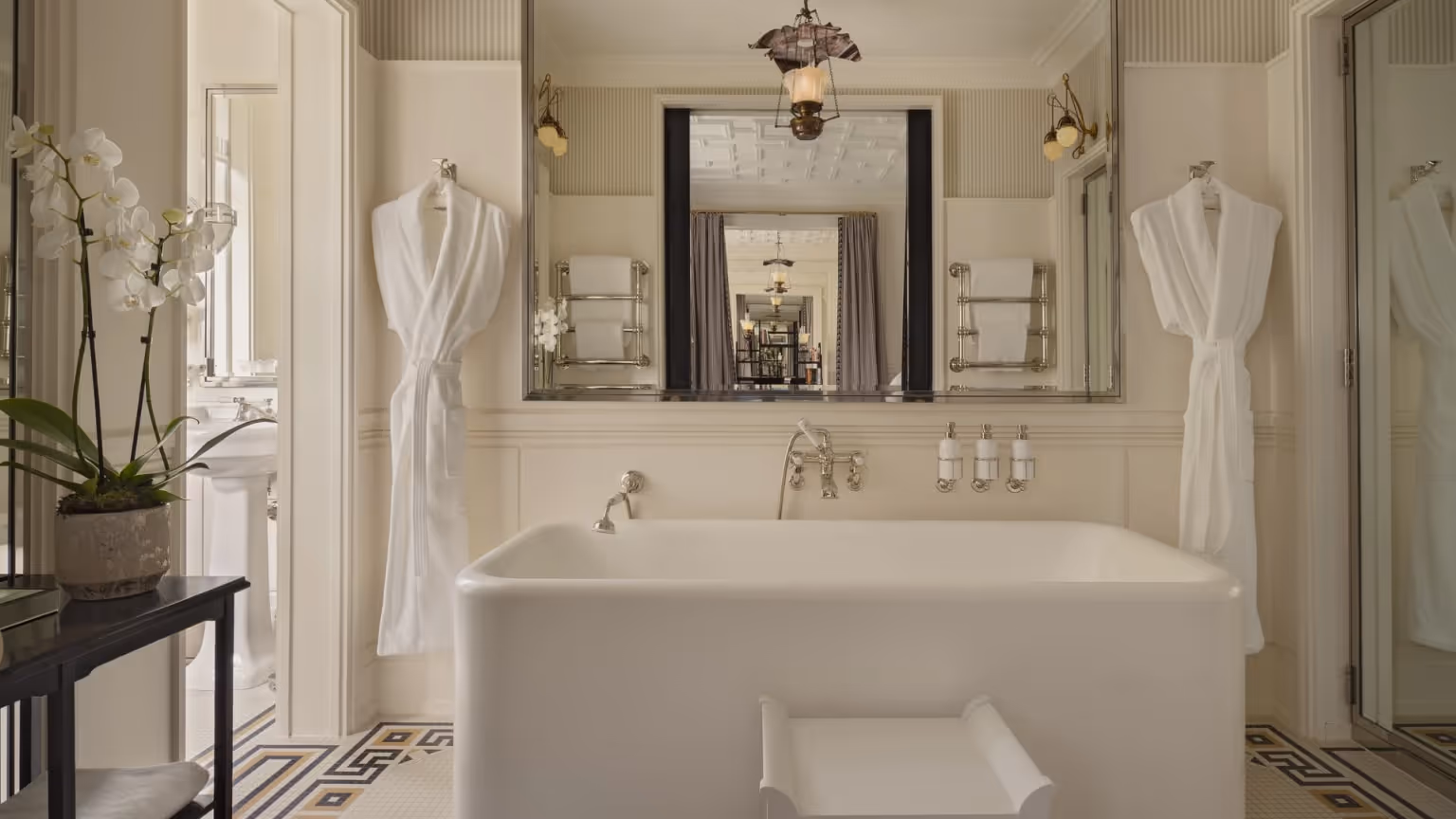 ALT:
White freestanding bathtub in a bright bathroom with mirrors, robes, and patterned floor tiles.