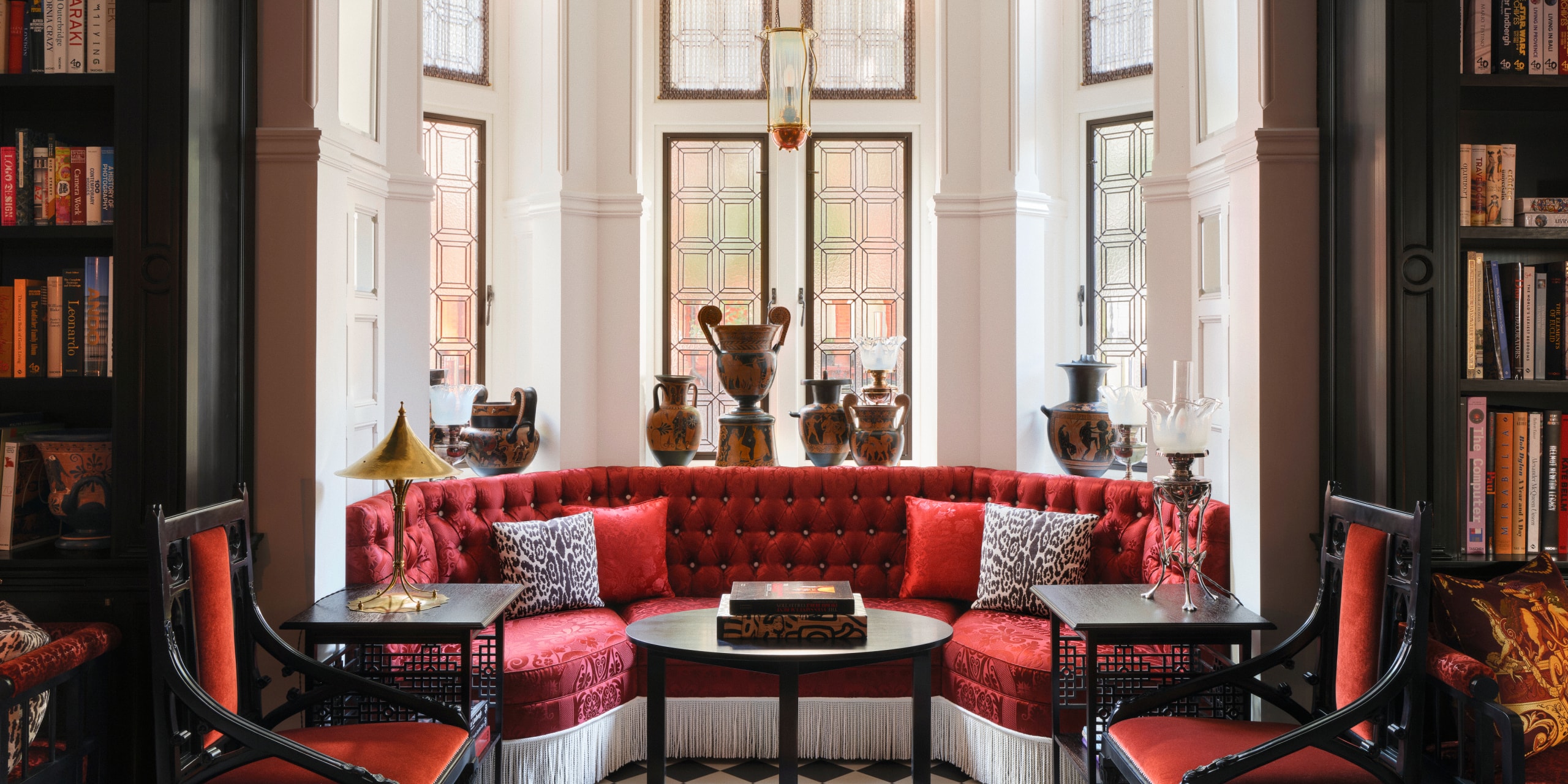 ALT: Window seating area in the lobby of At Sloane in London, featuring a red curved sofa, bookshelves, decorative objects and a traditional hotel lounge setting.