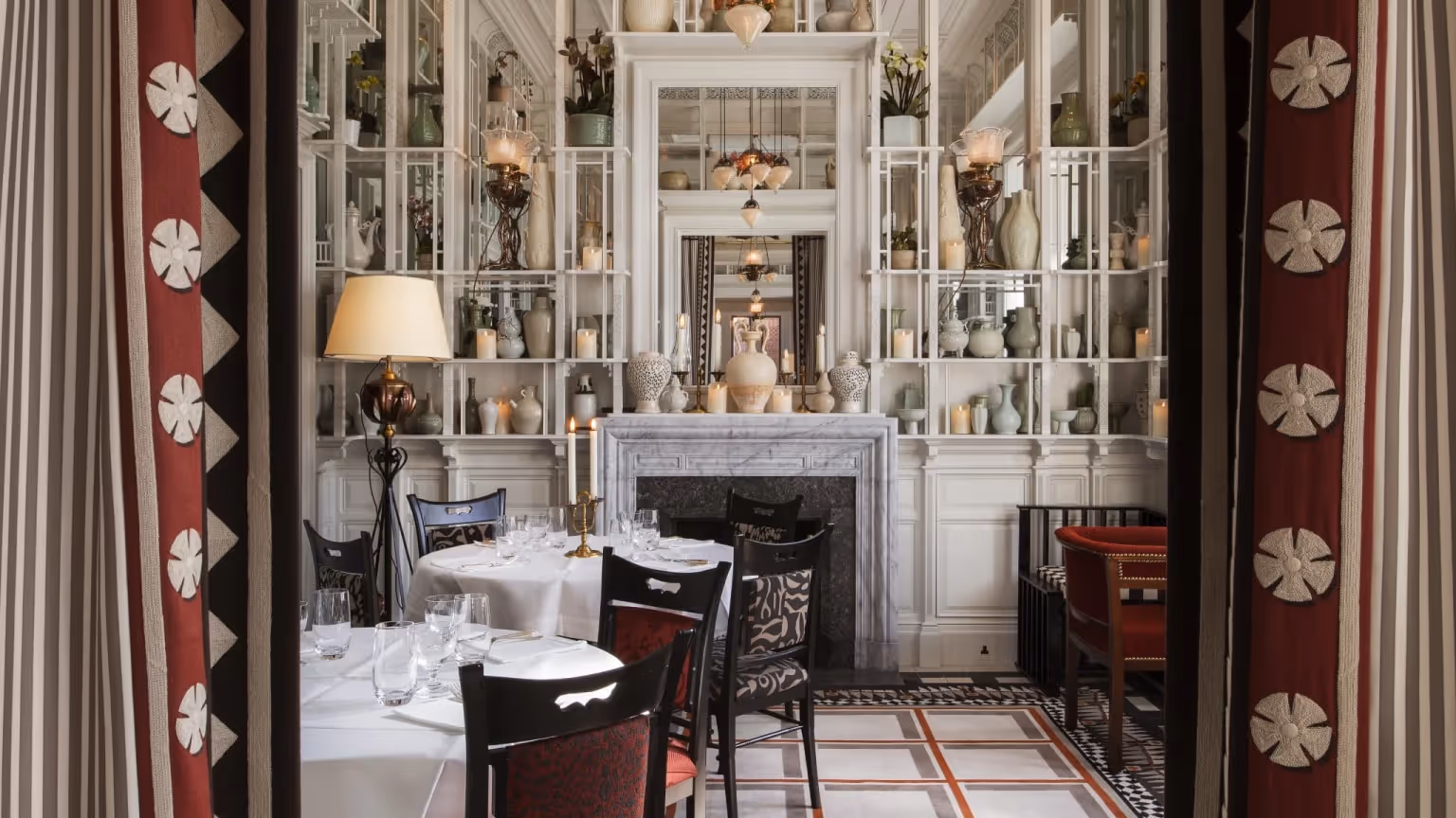 Top-floor dining room At Sloane in London with white built-in shelving, decorative objects, a fireplace, chandeliers, and red patterned curtains framing the space.
