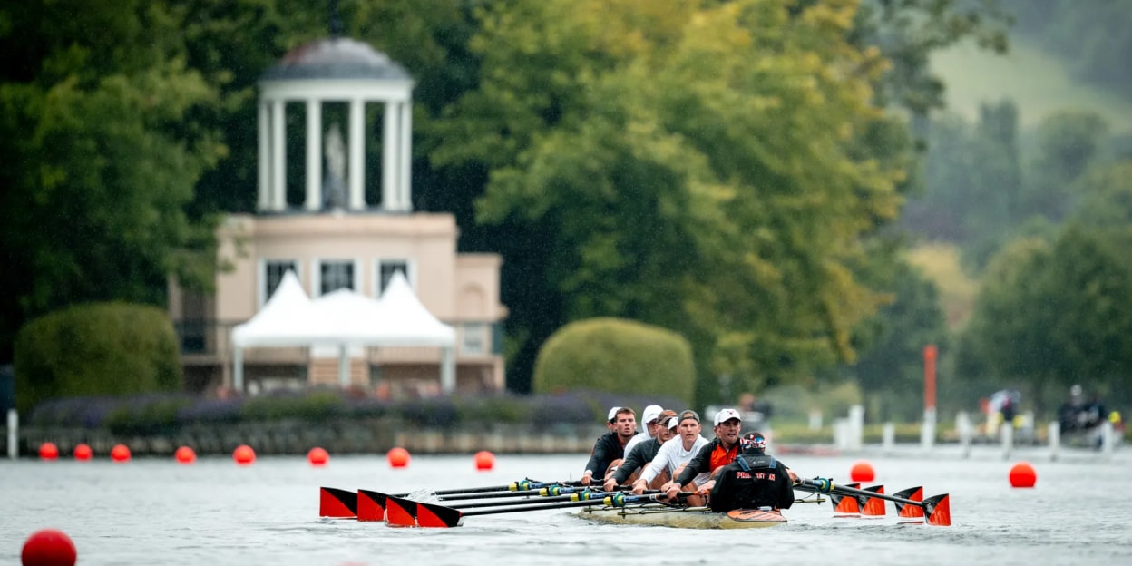 Rowing team racing on the River Thames with spectators and a pavilion visible on the riverbank in Henley, England.