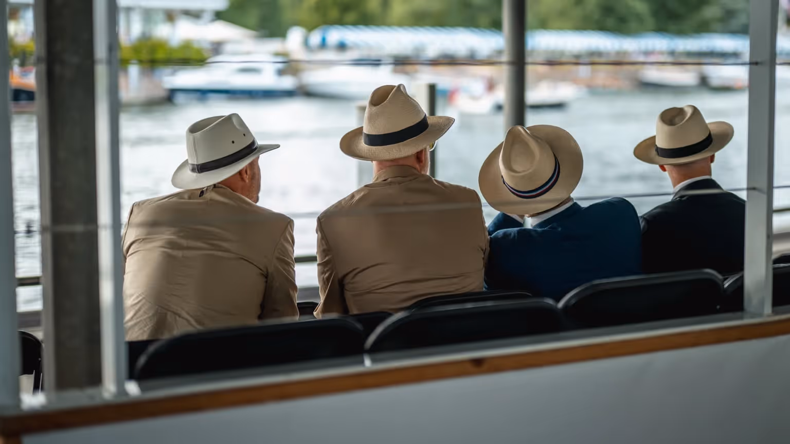 Four men wearing light-coloured suits and Panama hats sit facing the River Thames, watching boats pass during a summer event in Henley, England.