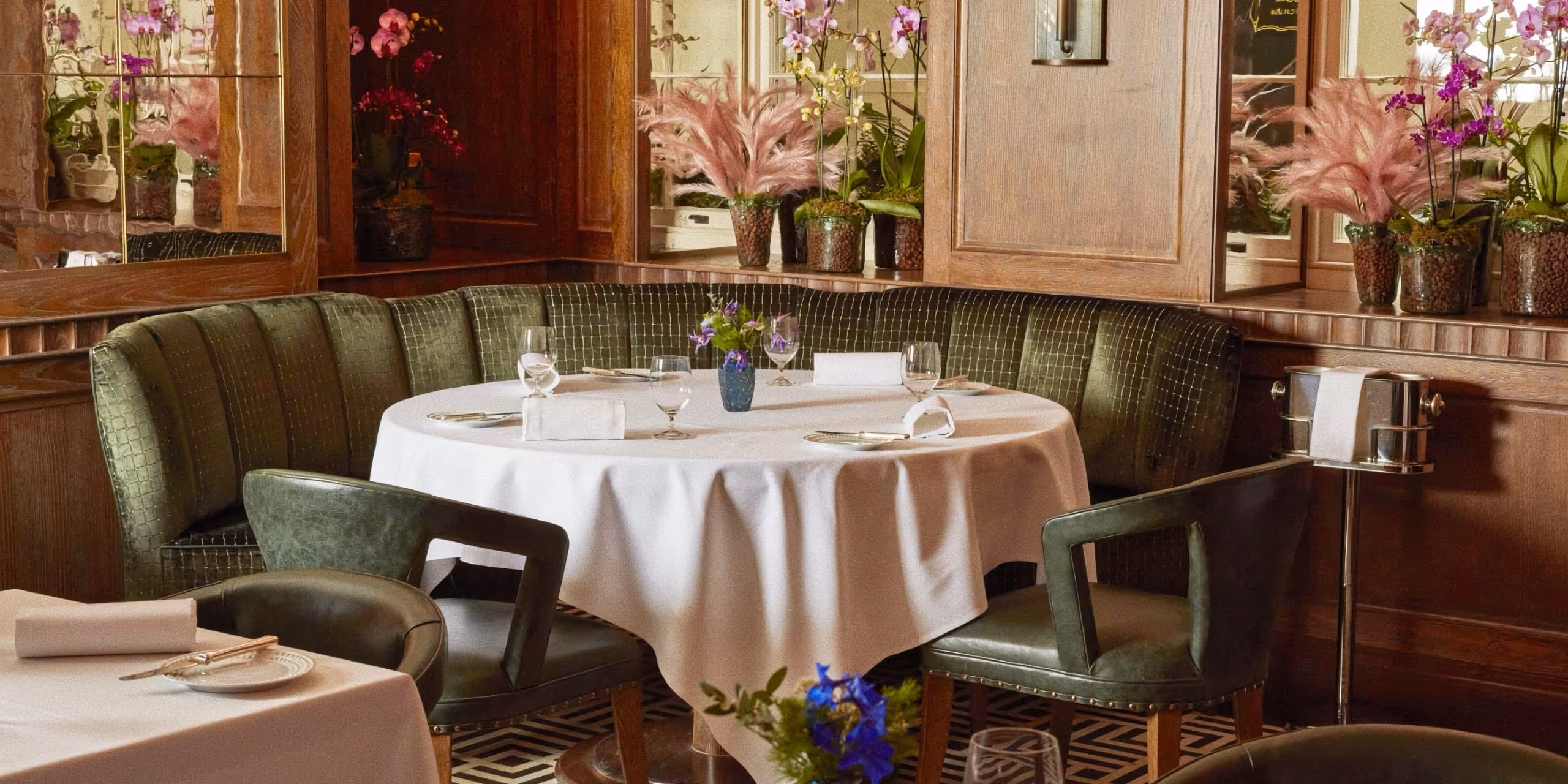 A luxury dining area at Ormer Mayfair featuring a curved green velvet booth, white linen tablecloths, and pink floral decorations in a wood-paneled room.