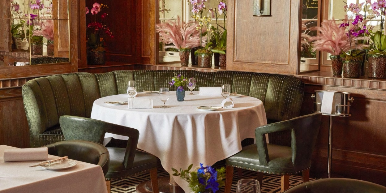 A luxury dining area at Ormer Mayfair featuring a curved green velvet booth, white linen tablecloths, and pink floral decorations in a wood-paneled room.