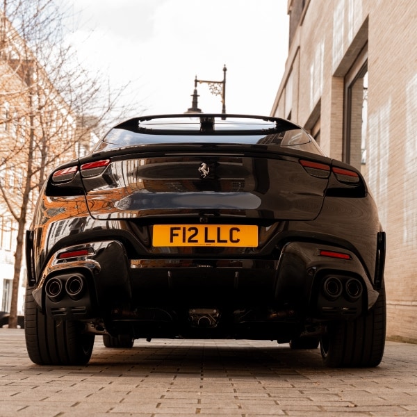 A powerful low-angle rear view of the black Ferrari Purosangue with custom F12 LLC plates, featuring quad-exhaust surrounds and aggressive aerodynamic lines on a London stone-paved street.