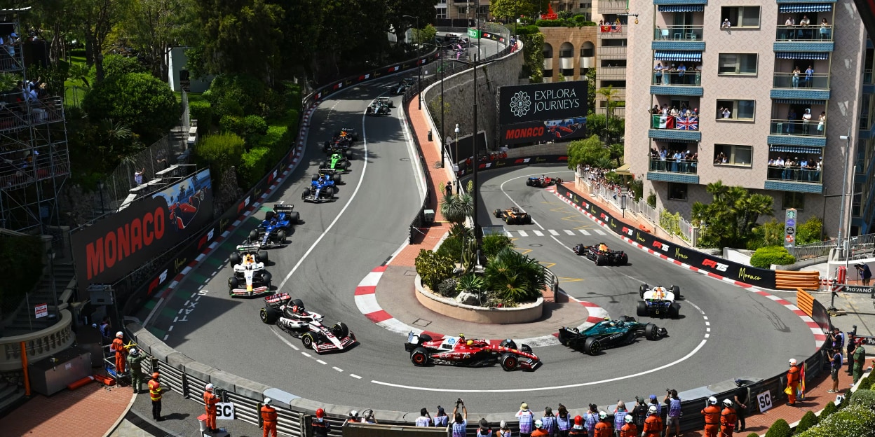 Several Formula 1 cars racing through the Fairmont Hairpin corner on the Monaco street circuit surrounded by buildings and spectators.