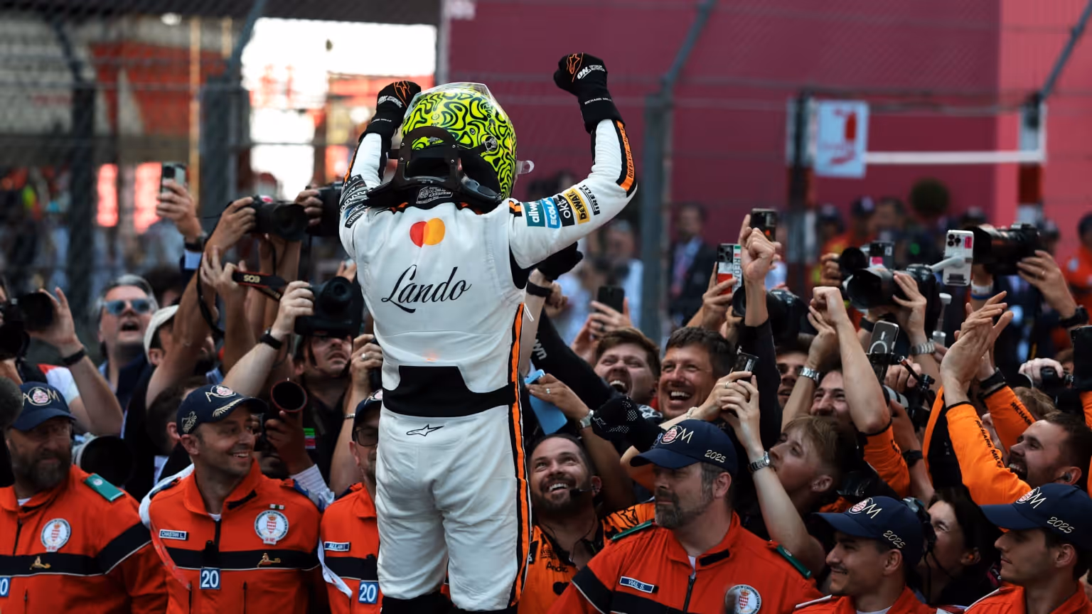A Formula 1 driver in racing gear raising his arms while surrounded by team members and photographers during a Monaco Grand Prix celebration.