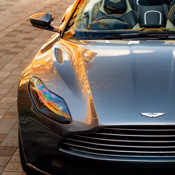 A close-up front view of the grey Aston Martin DB11 Volante showing the sculpted bonnet, LED headlight design, and iconic front grille in warm evening light.