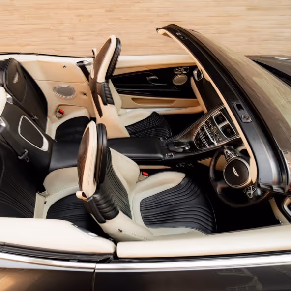 An elevated interior view of the Aston Martin DB11 Volante with the roof retracted, showcasing the luxury leather seats, center console, infotainment system, and elegant convertible cabin layout.