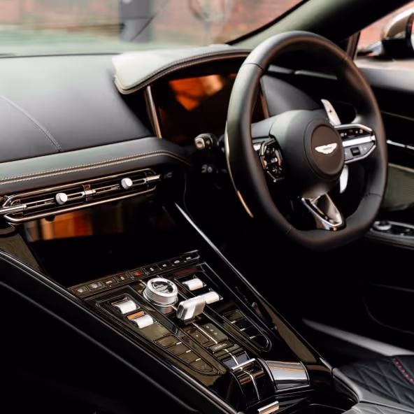 An interior view of the Aston Martin DB12 Volante showcasing the leather-wrapped steering wheel, digital dashboard, glossy center console, and handcrafted luxury cabin materials.