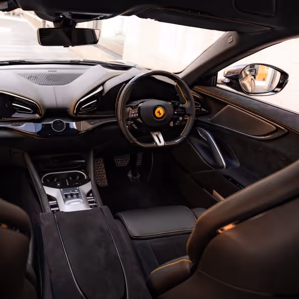 A wide interior view of the Ferrari Purosangue cockpit showcasing the dual-cockpit design, carbon fiber accents, and yellow contrast stitching on black leather.